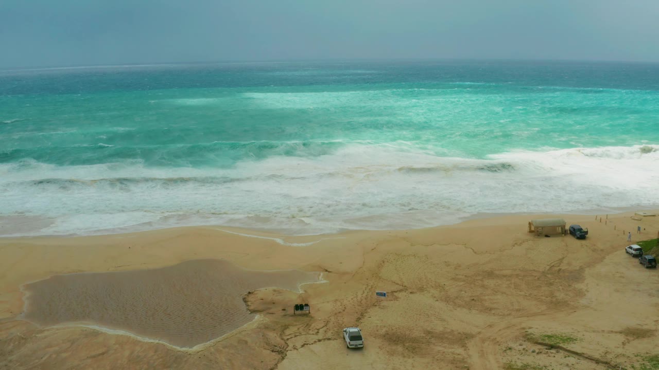 At Dreams Resort Los Cabos, foamy surf spreads across the middle of the frame while tan sand, a shallow pool, parked vehicles, and a small structure remain visible with no visible people.