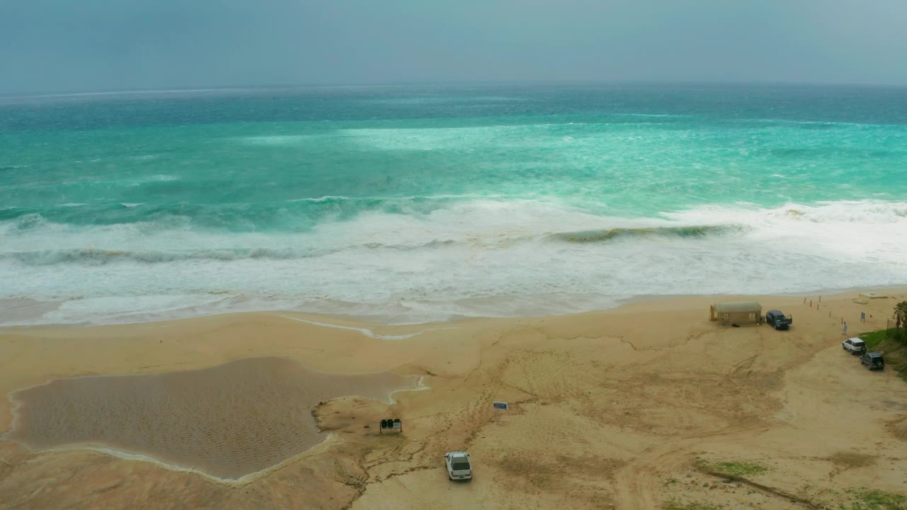 Aerial beach view with surf, tidal water, and vehicles parked on sand.