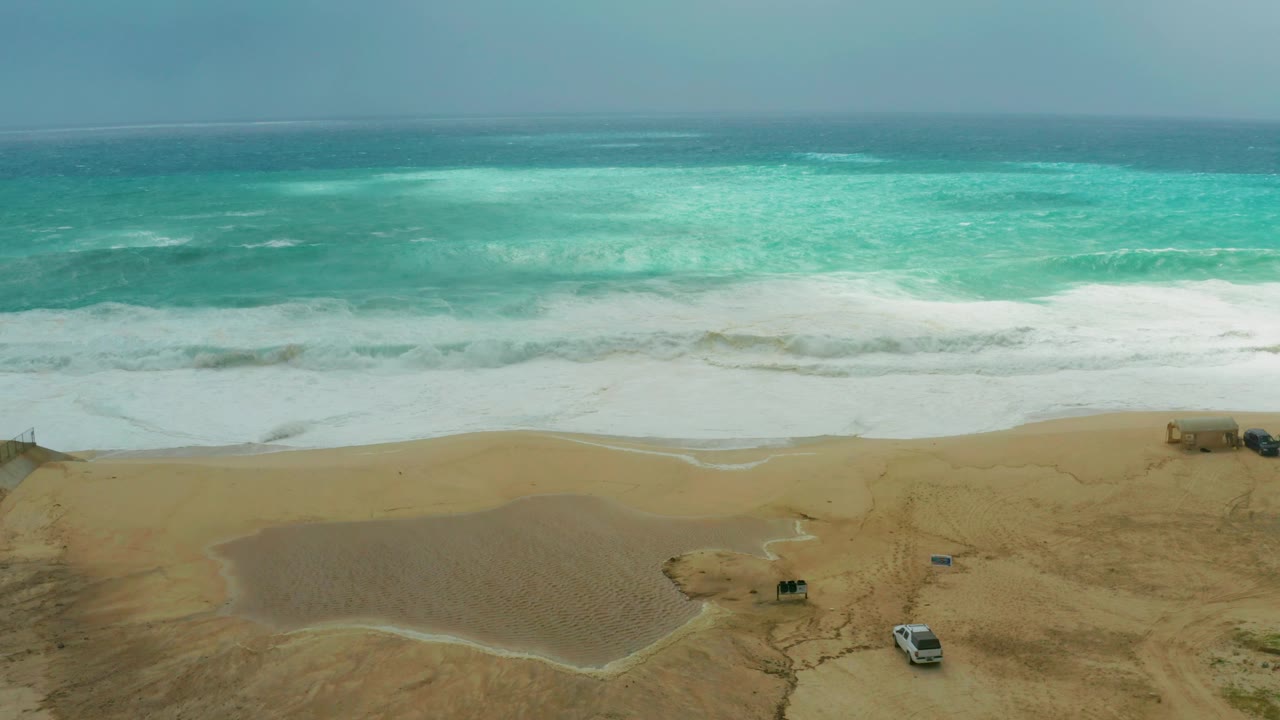 Wide elevated view at Dreams Resort Los Cabos, Mexico, showing turquoise surf, white foam, sandy beach, parked shore vehicles, and no visible people at the start of the clip.