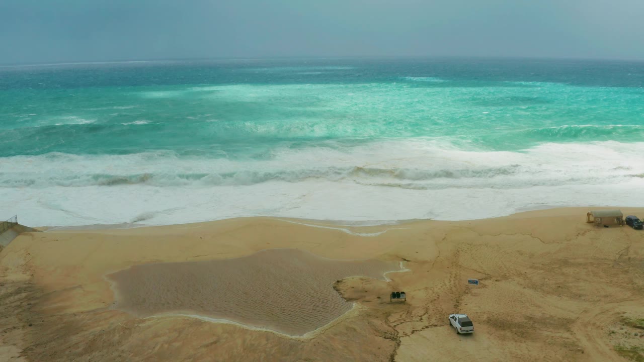 Foamy surf rolls toward a sandy shore with small parked vehicles.