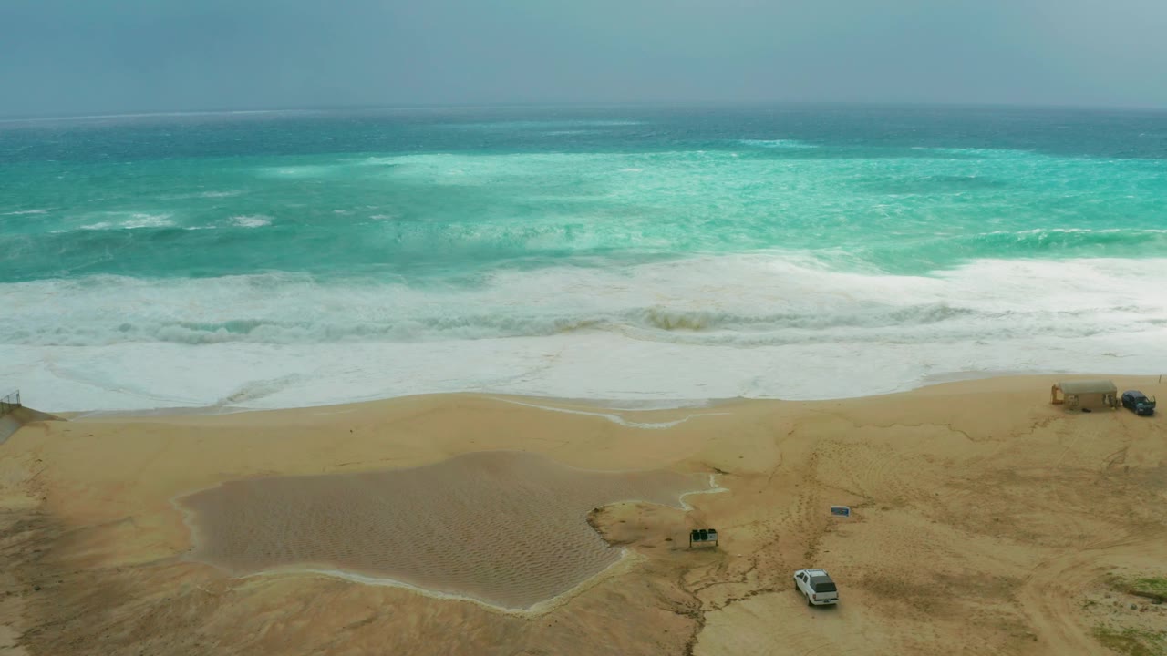 Aerial-style view of Dreams Resort Los Cabos beach where choppy turquoise water breaks into white foam above tan sand, with parked vehicles visible and no visible people.