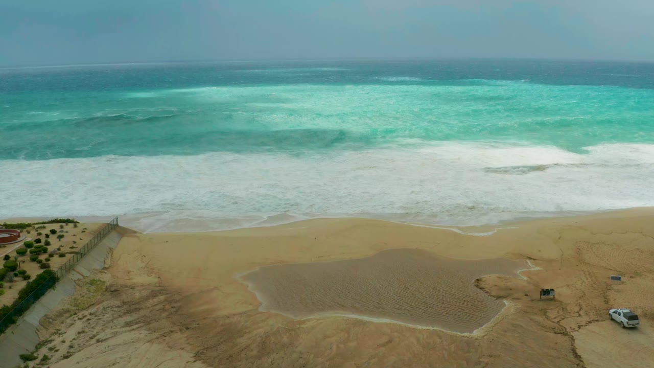 High aerial view of the beach at Dreams Resort Los Cabos, with turquoise surf rolling toward wide sand, a small vehicle near the lower right, and no visible people.