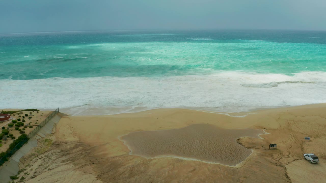Wide aerial view of the Dreams Resort Los Cabos shoreline, with bright turquoise water, a broad white wave band, open sand, a small vehicle farther left than before, and no visible people.