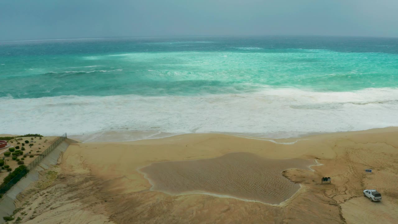 Aerial beach frame at Dreams Resort Los Cabos with foamy surf advancing toward shore, textured sand below, the vehicle still visible near the right side, and no visible people.