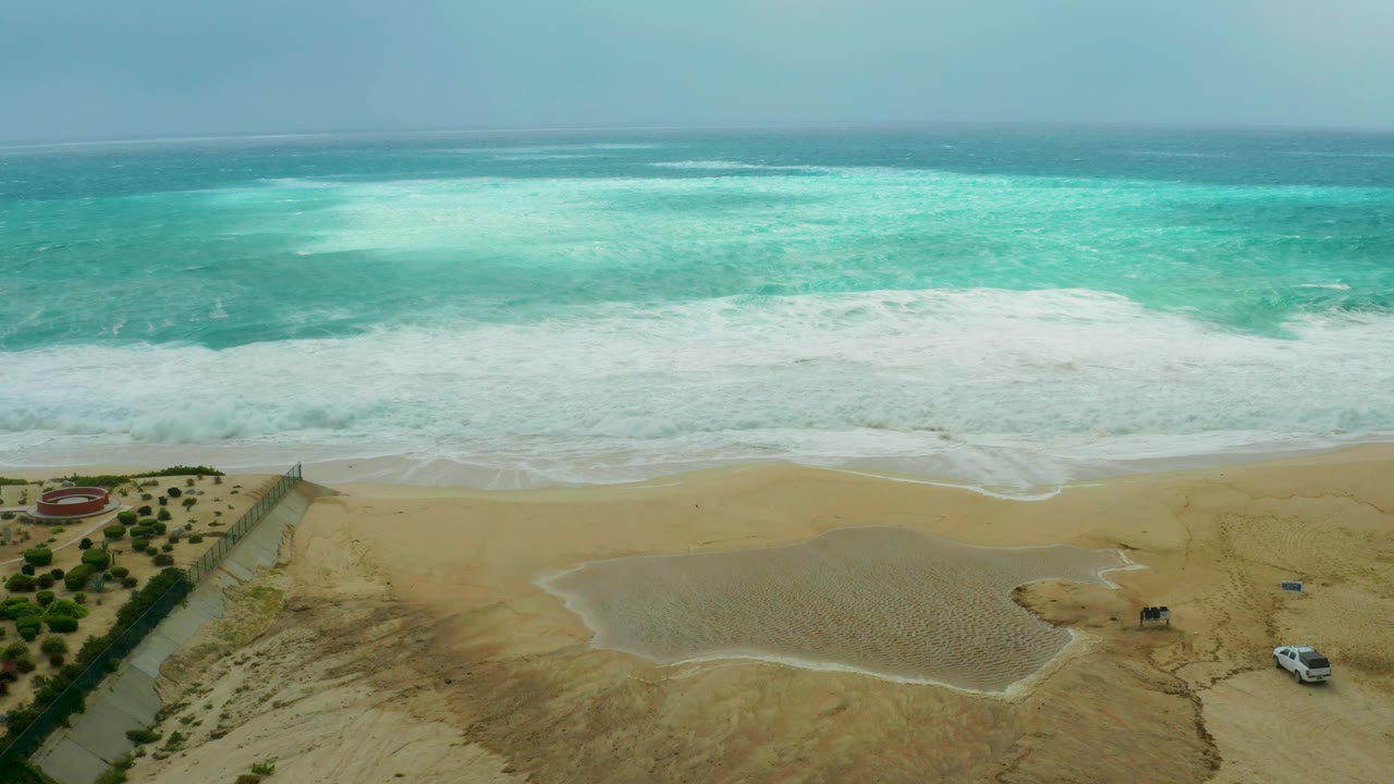 Aerial frame at Dreams Resort Los Cabos showing turquoise ocean, white surf, wet sandy beach, a shallow pond, sparse vegetation, a road edge, and no visible people.