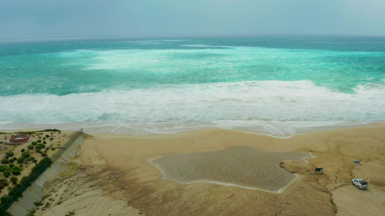 Dreams Resort Los Cabos beach from above with turquoise water darkening toward the horizon, a broad band of white surf, ponded sand, vegetation patches, a road, and no visible people.