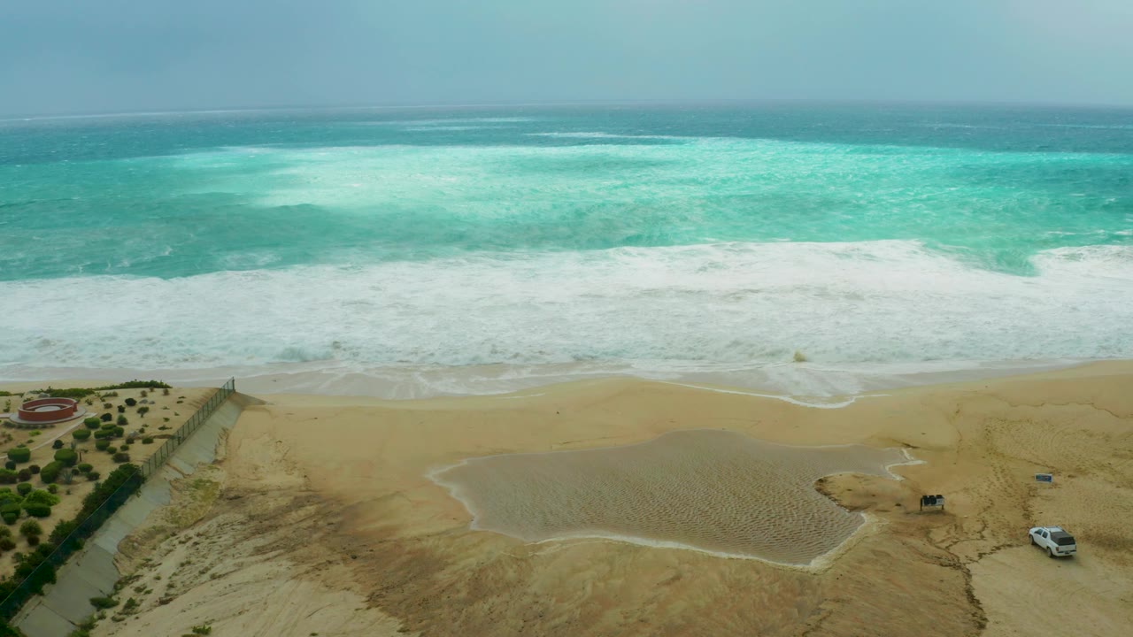 Wide aerial view of surf breaking onto sand with a pond and small car near the lower right.