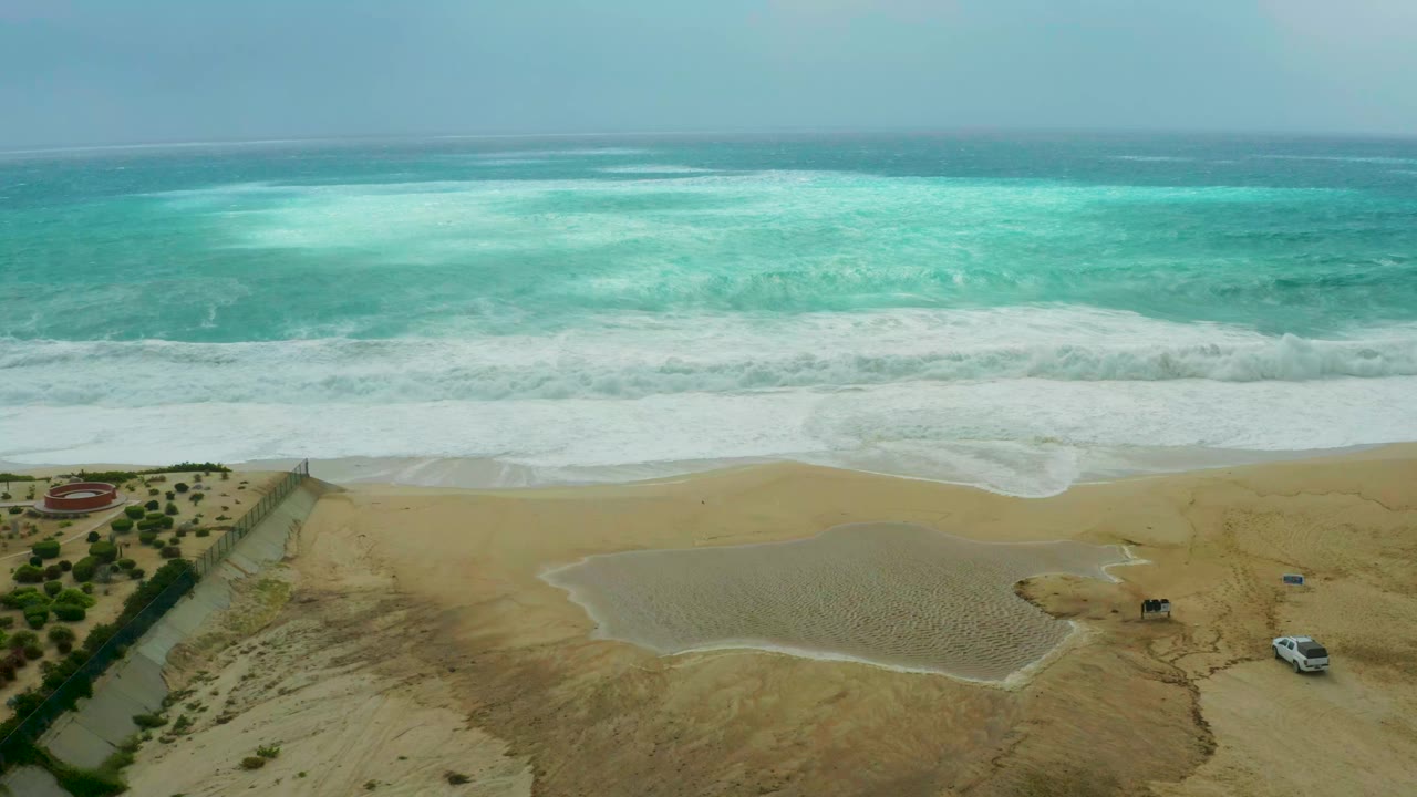 At 0:01, the Dreams Resort Los Cabos shoreline shows a broad band of white foam moving across sand, with the path, greenery, and white vehicle lower in frame and no visible people.