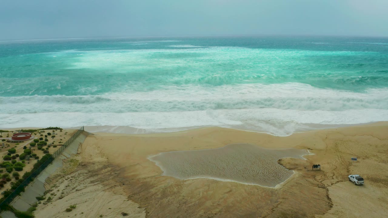 At 0:03, the surf line sits slightly lower across the Dreams Resort Los Cabos sand, with turquoise ocean beyond, a beach path at left, a white vehicle at lower right, and no visible people.