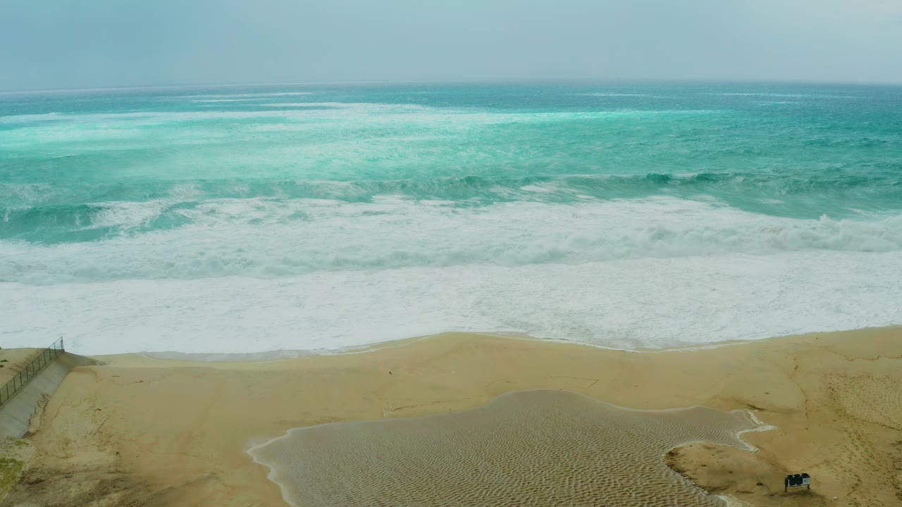 Aerial frame from Dreams Resort Los Cabos showing turquoise waves breaking onto sand, dune fencing at the lower left edge, small vehicles on the beach area, and no visible people.