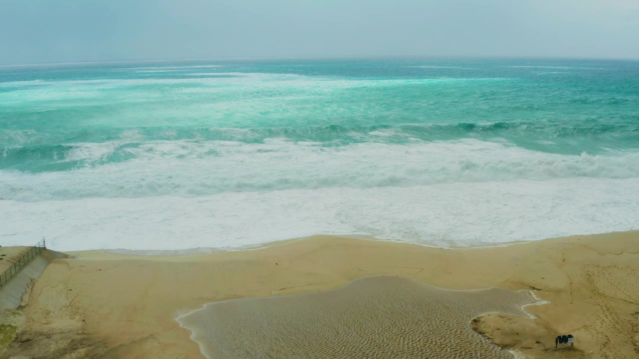 At 0:01, the Los Cabos beach view holds a broad band of white foam, tan sand, access fencing near the lower left, a dark vehicle on the sand, and no visible people.