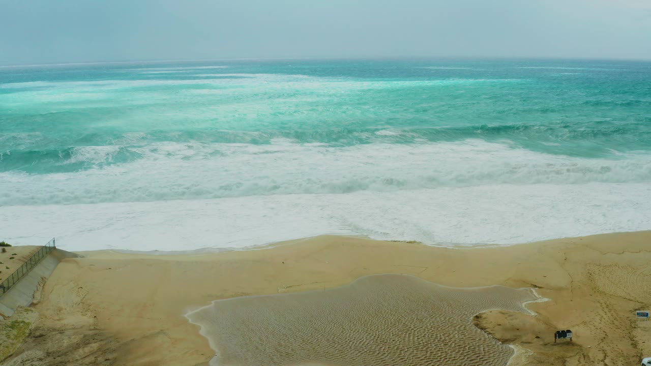 At 0:02, waves advance toward the shore at Dreams Resort Los Cabos while the lower beach shows fencing, vehicle tracks, small vehicles, and no visible people.