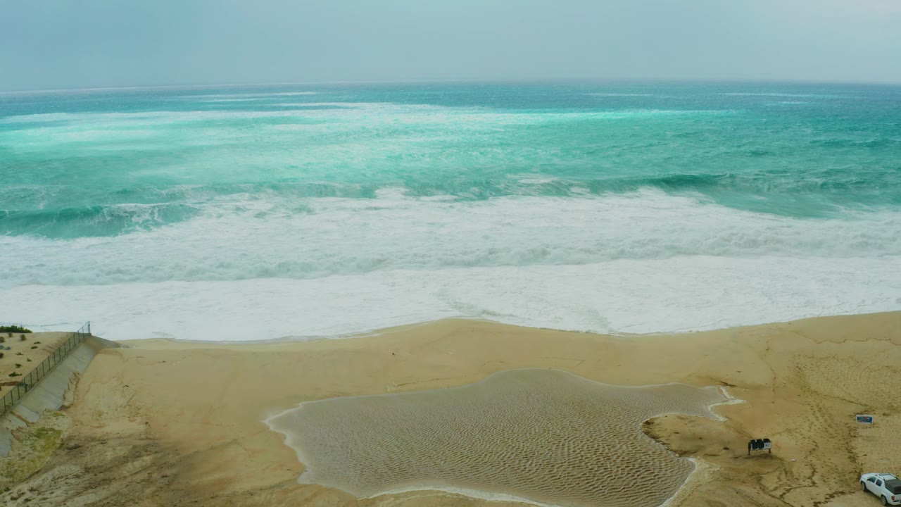 At 0:03, the aerial view shows a white vehicle near the lower right edge, dune fencing at lower left, rolling surf beyond the sand, and no visible people.