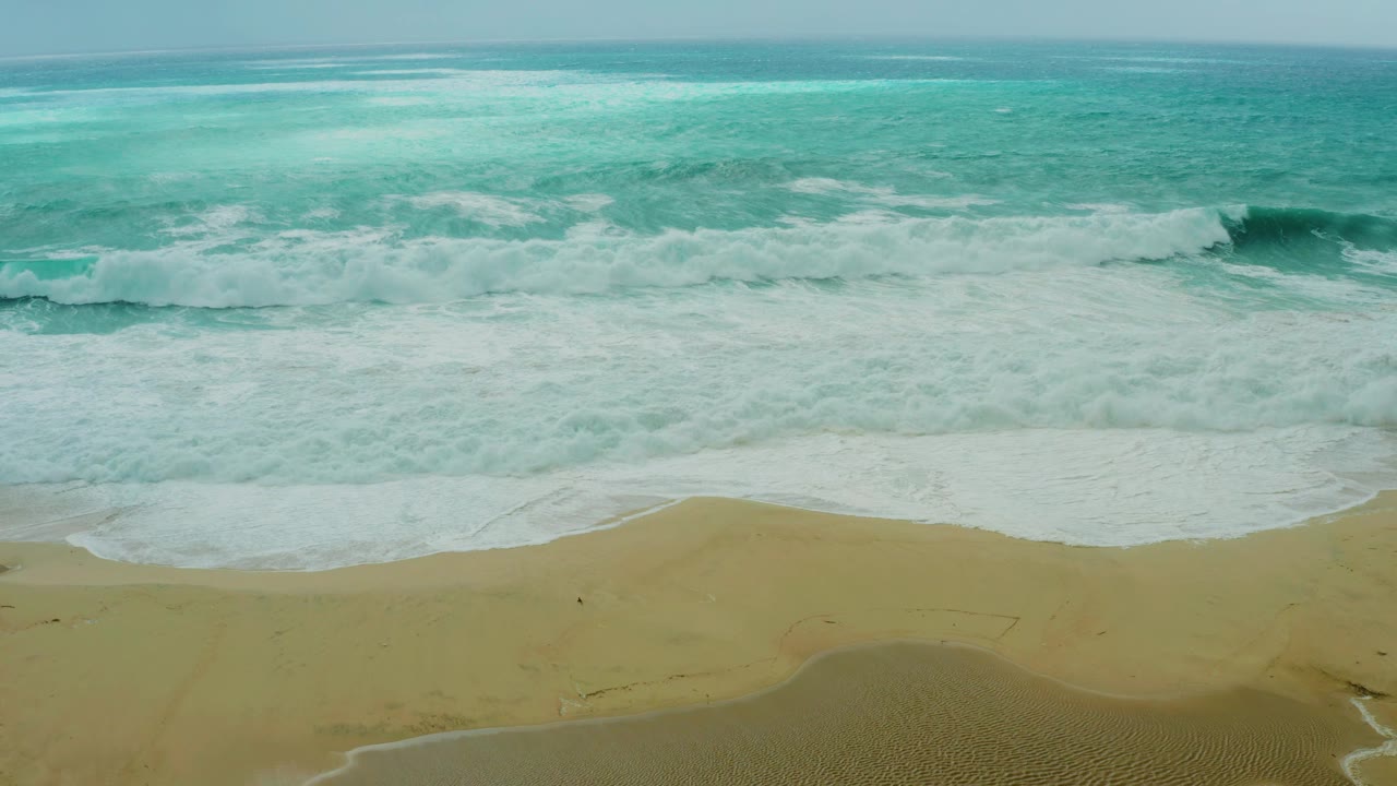 Elevated view of turquoise ocean waves breaking onto the sandy beach at Dreams Resort Los Cabos, with white foam across the shoreline and no visible people.