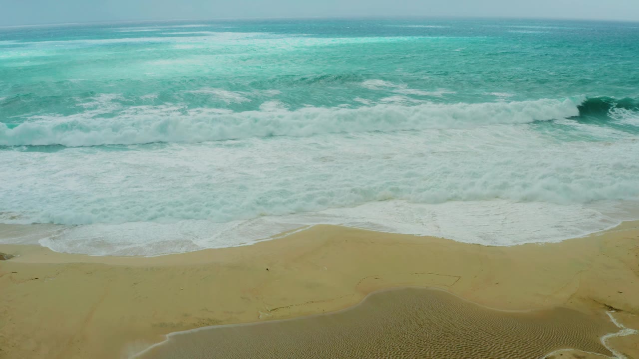 Foamy surf covers the center of the frame as turquoise water sits beyond the break and tan beach remains clear below at Dreams Resort Los Cabos, with no visible people.