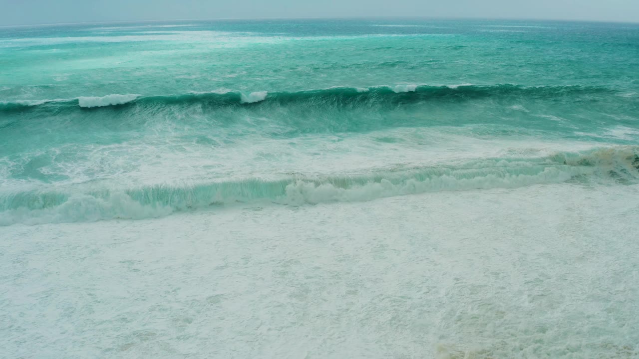 Aerial shoreline frame at Dreams Resort Los Cabos with whitewater widening across the center and pale sand partly covered by foam, with no visible people.