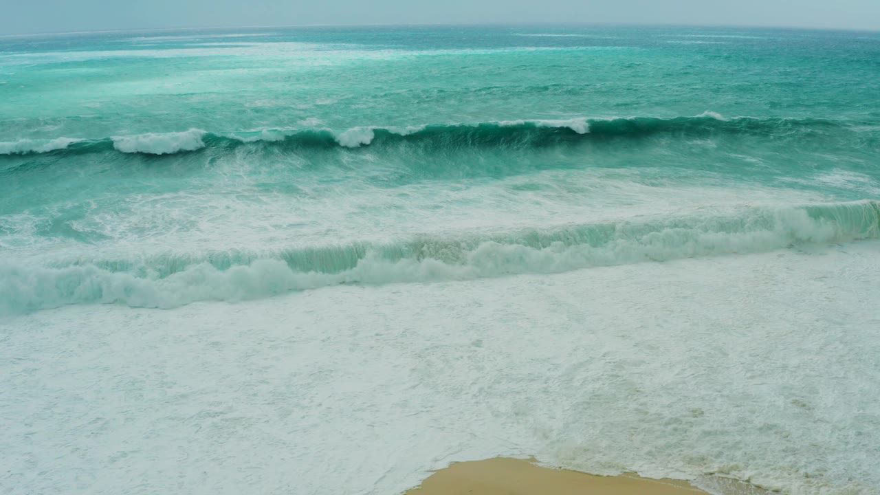 Aerial view of the Los Cabos beach as a foamy wave washes farther down the frame toward tan sand, with no visible people or structures.