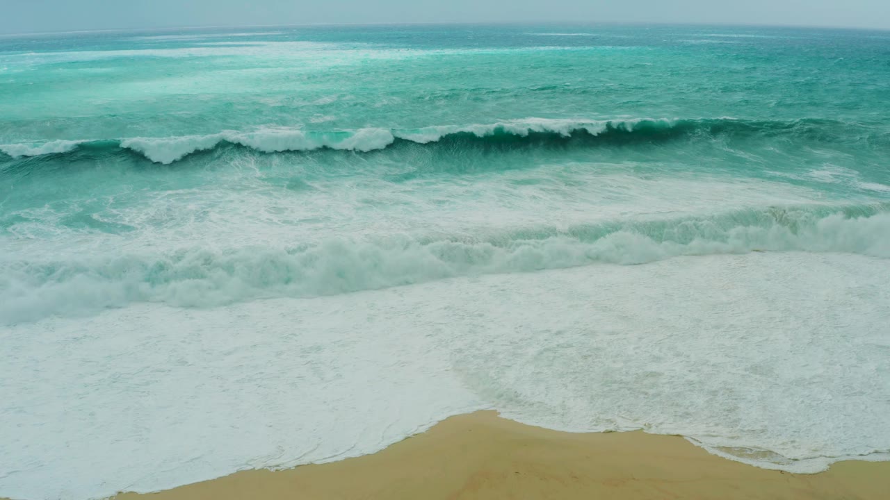 Wide aerial composition at Dreams Resort Los Cabos with a long horizontal surf line, turquoise sea beyond it, and empty sand below, with no visible people.