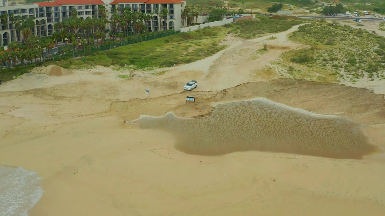 Aerial frame at Dreams Resort Los Cabos showing a broad tidal pool on pale sand, surf at the lower left edge, dunes, palms, resort buildings, and no visible people.