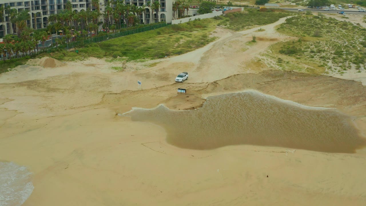 Aerial frame from Dreams Resort Los Cabos showing the tidal pool shifted slightly lower in the composition, with dunes, a path, a white vehicle, palms, and no visible people.