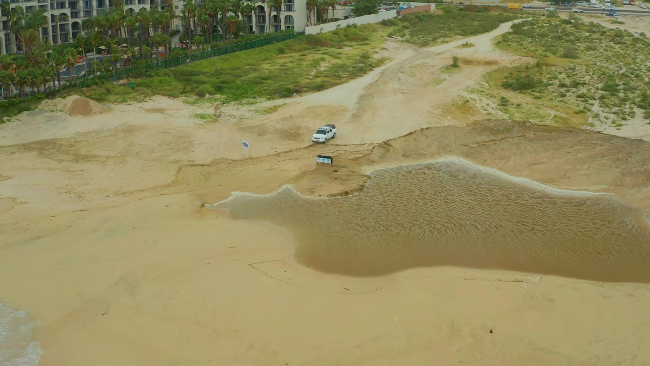 Aerial beach scene with shallow water pool, dunes, palms, and vehicle.