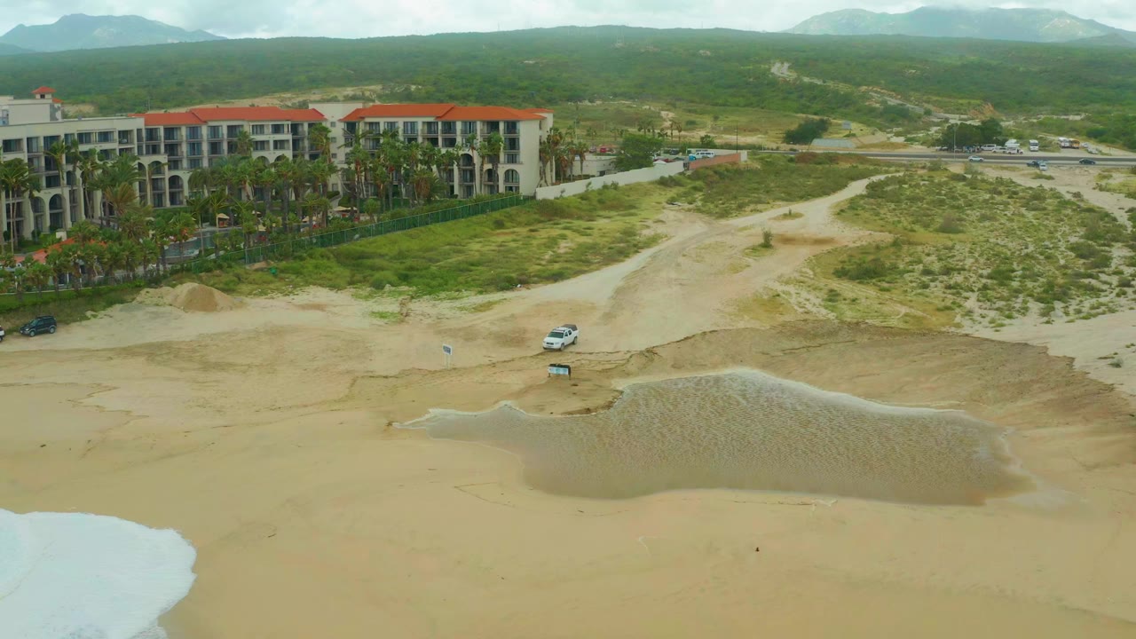 At 0:01, the Los Cabos beach remains in a high aerial composition, with shallow surf at the lower left, the white vehicle near the wet sand, resort buildings and palms at left, green hills in back, and no visible people.