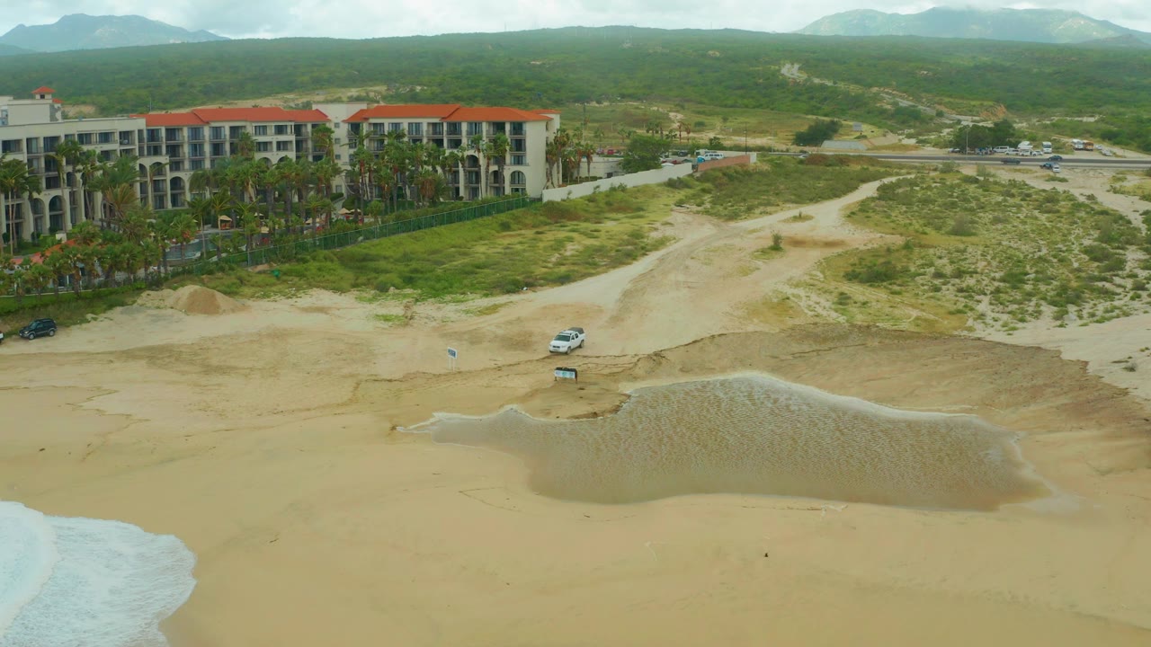 Aerial resort beach scene with surf at lower left, parked white vehicle, sand, palms, buildings, and hills.