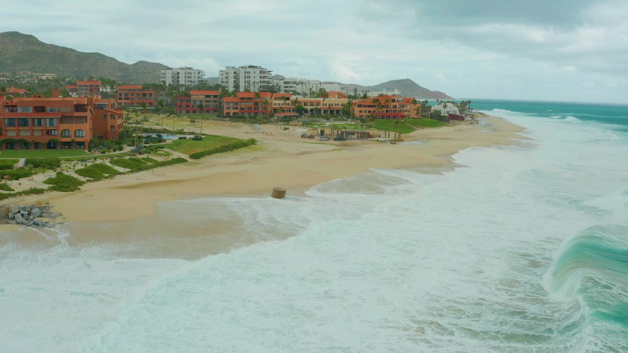 Opening frame of Dreams Resort Los Cabos beach with white surf spreading over sand, red-orange coastal buildings, cloudy sky, hills, and no visible people.