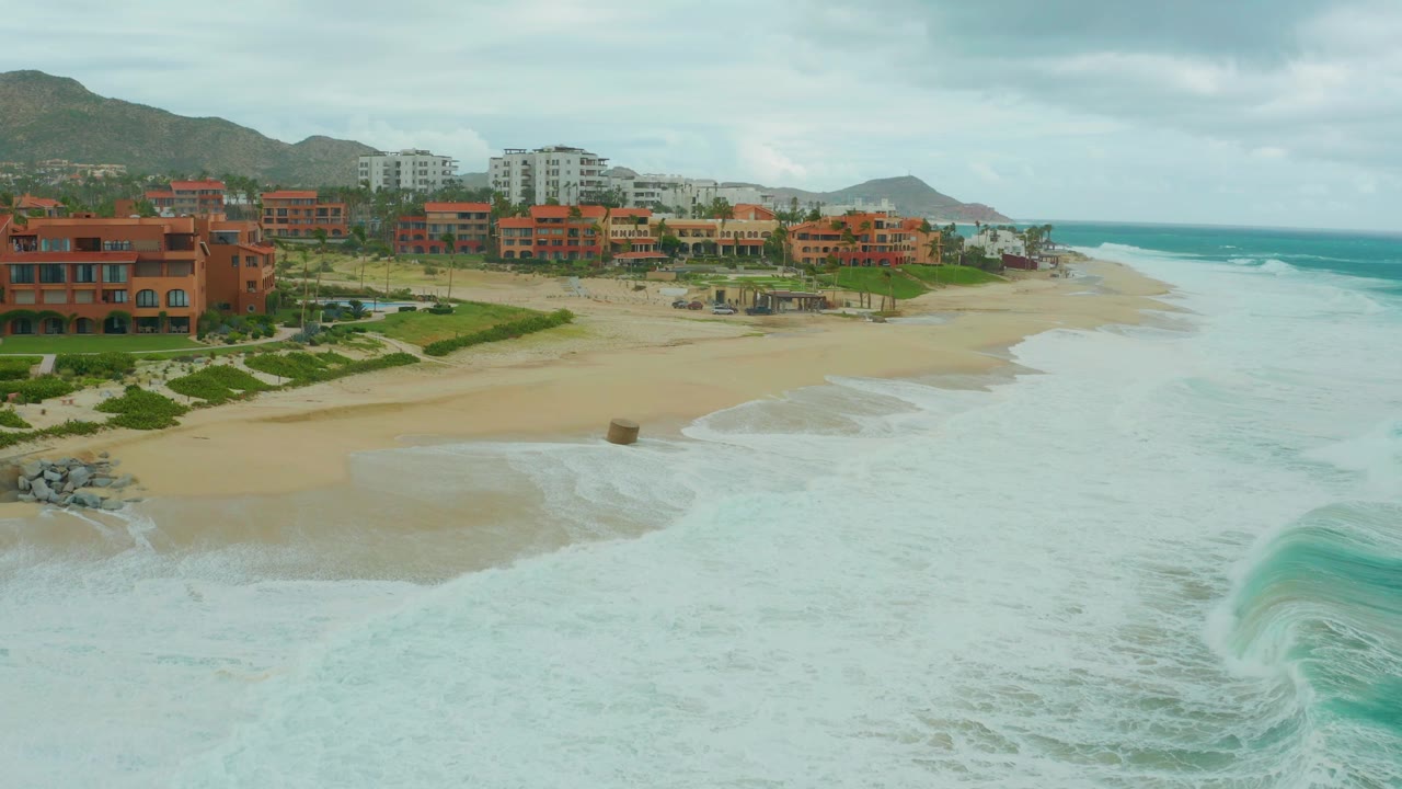 Foamy surf advances across the beach in front of resort buildings and hills.
