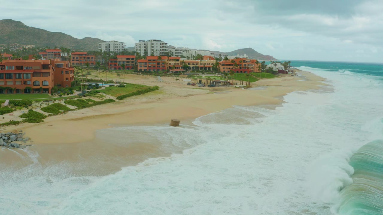 A later view of Dreams Resort Los Cabos beach where dense white surf occupies more of the right side, with sand, coastal buildings, greenery, hills, and no visible people.