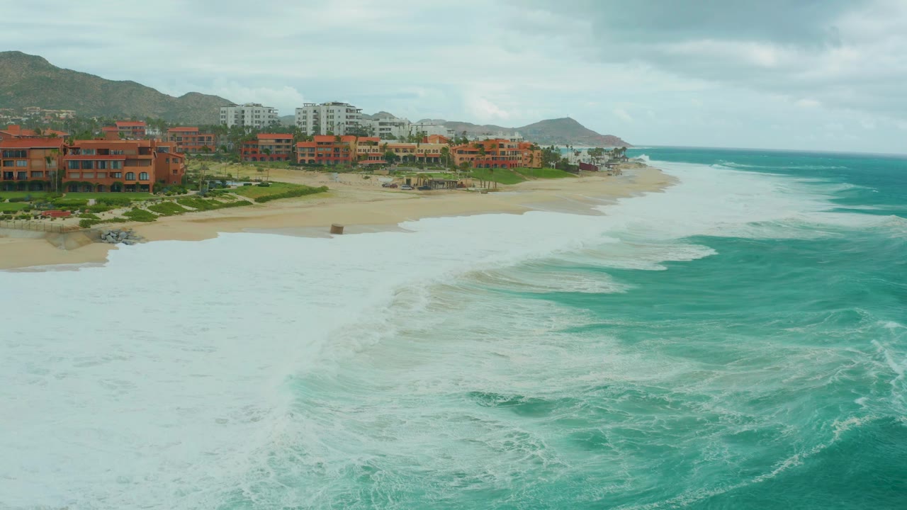 Aerial view at Dreams Resort Los Cabos with a broad band of whitewater moving across the near water, red-orange beachfront buildings on the sand, distant mountains, cloud cover, and no visible people.