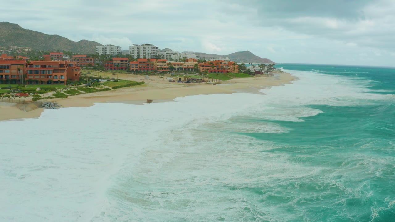 Drone-height frame over Dreams Resort Los Cabos with a stronger curl of turquoise wave at right, thick foam across the foreground, sandy beach in the center, resort buildings behind it, and no visible people.