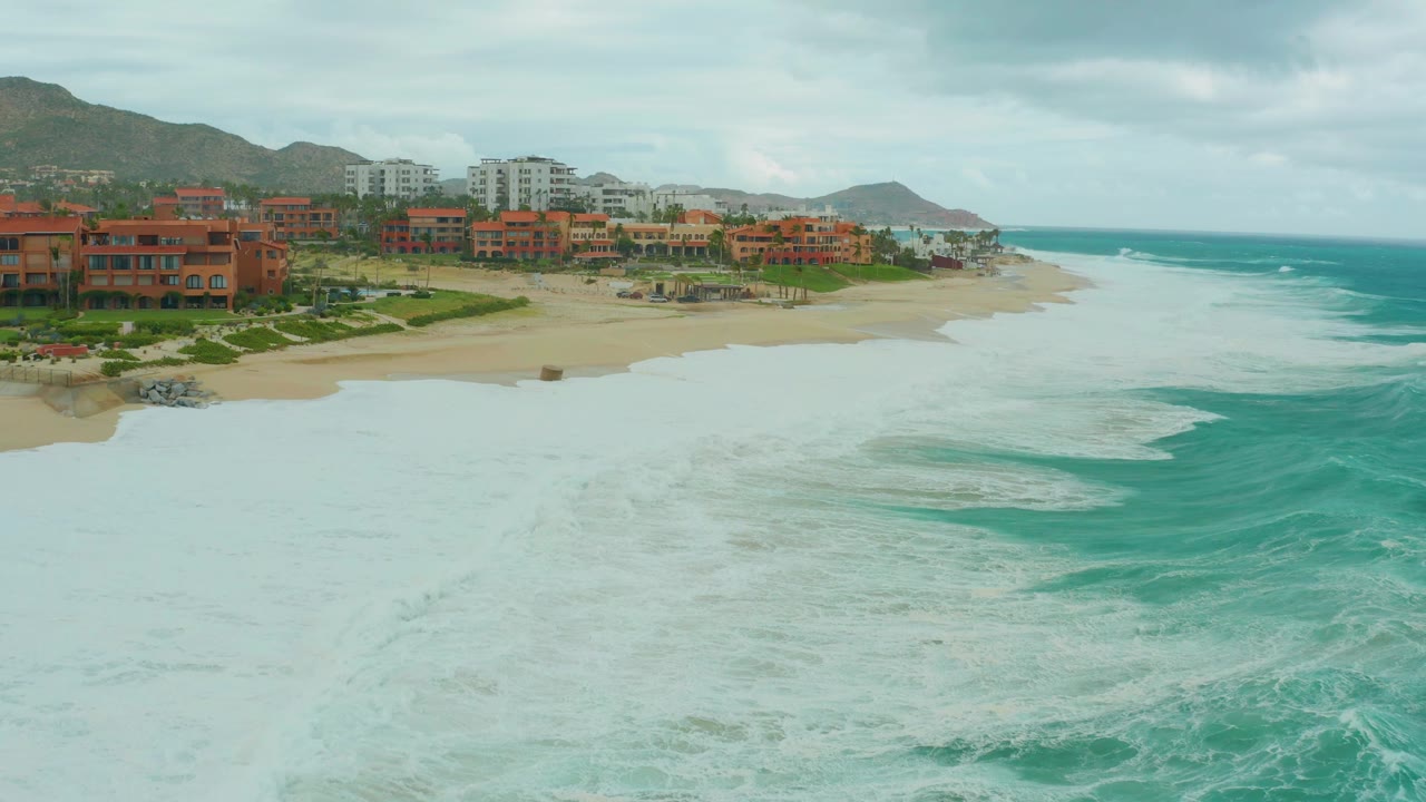 Aerial frame at Dreams Resort Los Cabos where pale foam covers most of the foreground, the sandy beach arcs behind it, resort buildings sit along the left side, mountains line the background, and no visible people appear.