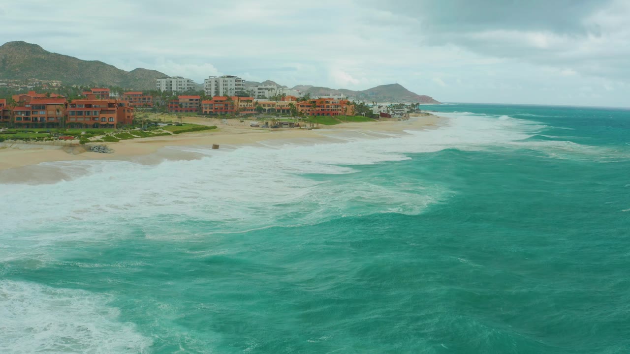 Opening aerial frame at Dreams Los Cabos, Baja California Sur, showing turquoise ocean and white foam in the foreground, beachfront buildings and hills beyond, with no visible people.
