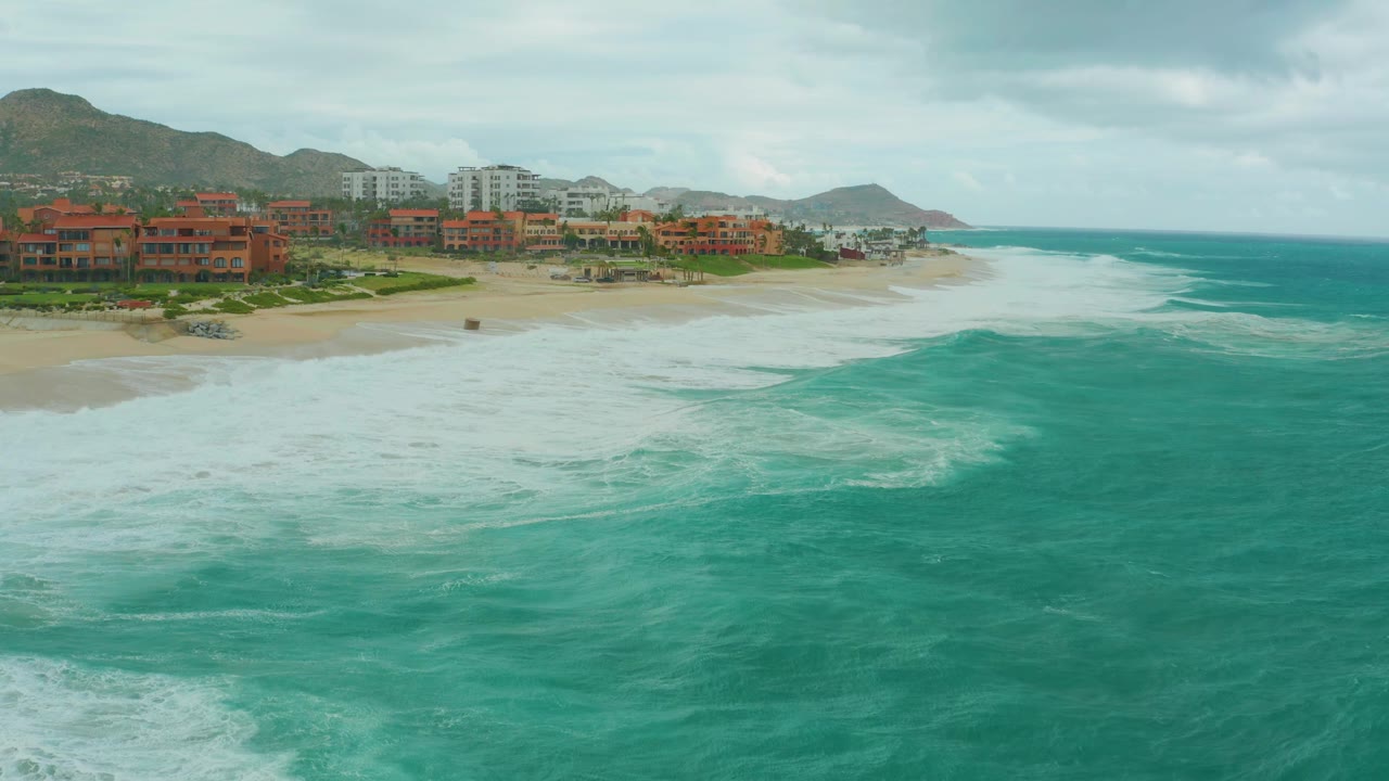 Aerial view at Dreams Los Cabos showing turquoise water curving into foamy surf near the beach, resort buildings along the left shoreline, mountains in back, and no visible people.