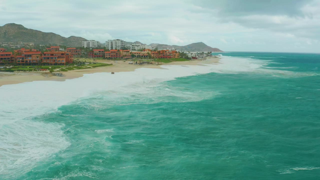 Aerial frame at Dreams Resort Los Cabos showing white wave wash spreading along the sand, red and white coastal buildings, distant hills, cloudy sky, and no visible people.