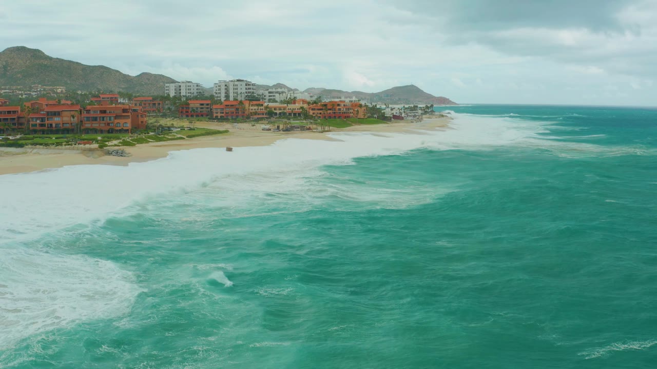 Aerial view at Dreams Resort Los Cabos with a curved patch of foam in turquoise water, sandy shoreline, clustered coastal buildings, mountain backdrop, gray clouds, and no visible people.