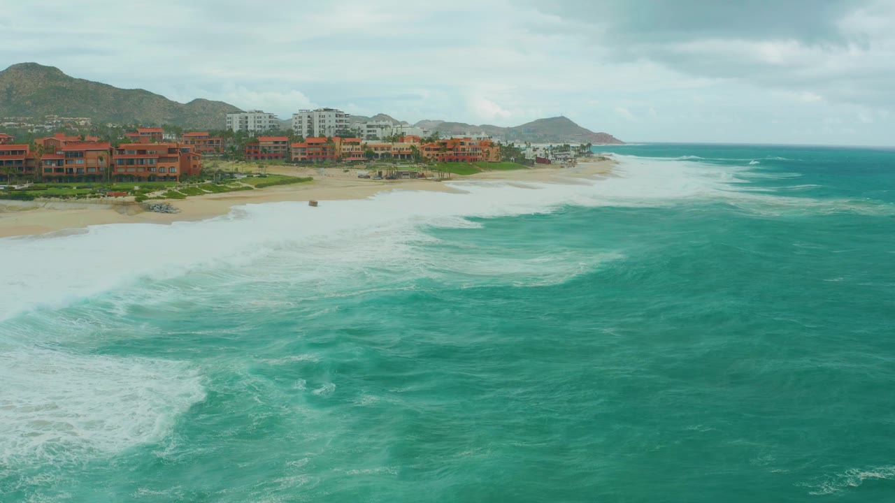 Mid-clip aerial frame at Dreams Resort Los Cabos with a broad white surf field near the sand, red coastal buildings, white buildings, mountains, overcast sky, and no visible people.