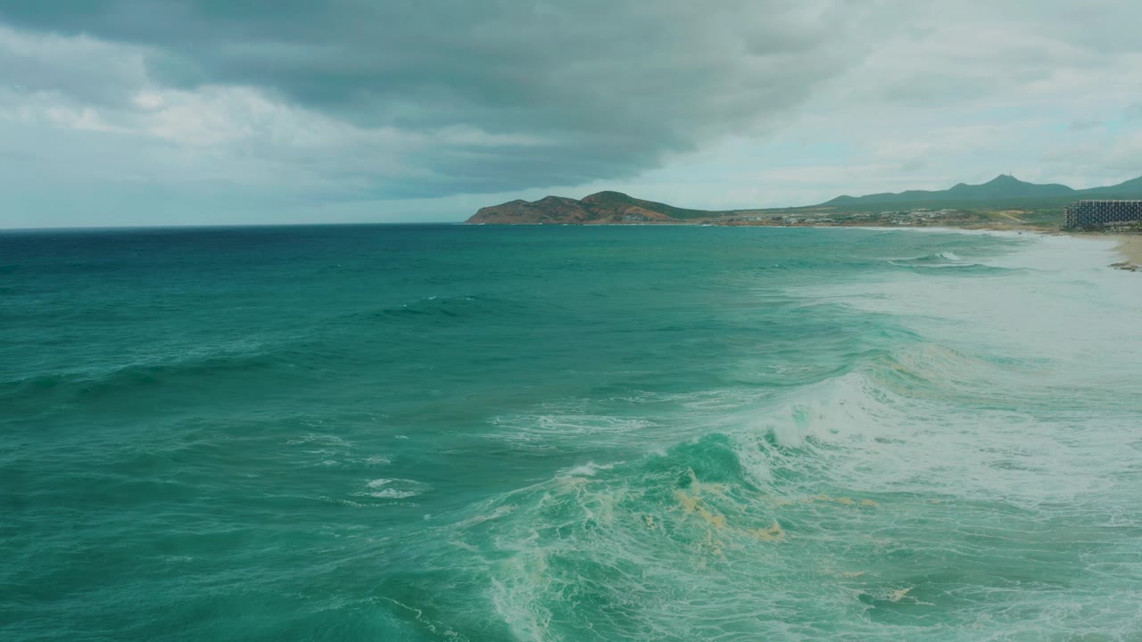Dreams Resort Los Cabos coastline with a broad band of sea foam over turquoise water, hills on the horizon, dark clouds above, and no visible people.
