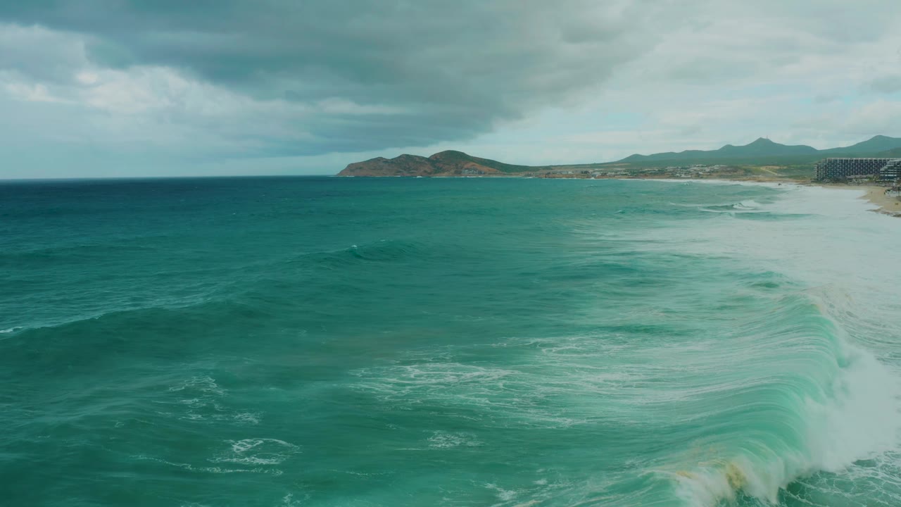 Dreams Resort Los Cabos aerial view with a swell forming in the darker water, white foam washing toward the beach, hills in the background, and no visible people.