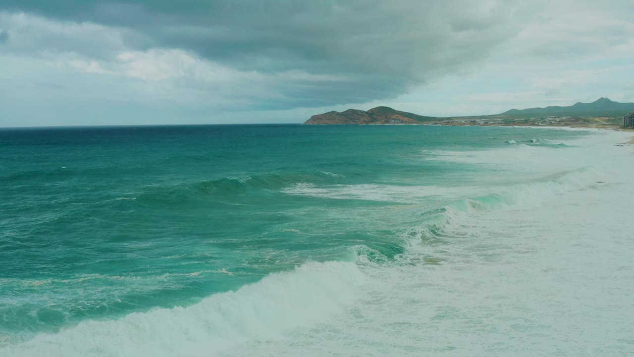 Turquoise ocean waves breaking near a sandy coast under cloudy sky.