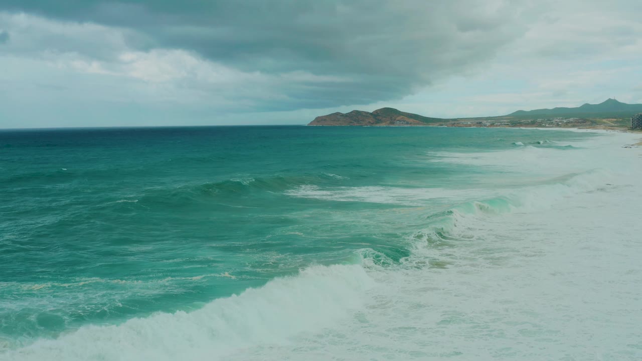 At 0:01, the Dreams Resort Los Cabos shoreline remains distant while white foam widens across the right side of the turquoise ocean, with no visible people.