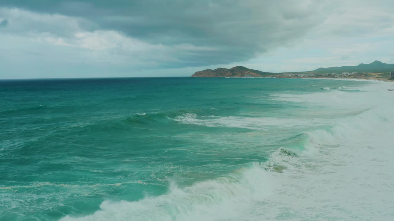 At 0:02, the wave face is more pronounced in the lower right while distant hills and the cloudy sky hold the background at Dreams Resort Los Cabos, with no visible people.