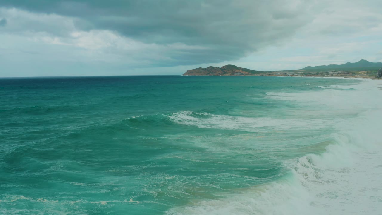 At 0:03, the frame shows a broader sheet of white surf across the midground water off Dreams Resort Los Cabos, with hills on the horizon and no visible people.