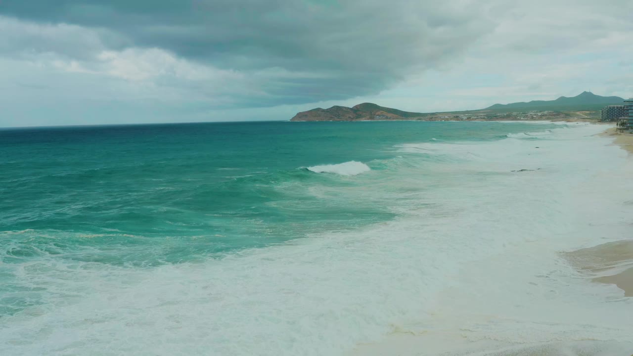 At 0:01, a foamy wave line advances across the shallow water near the sandy shore at Dreams Resort Los Cabos, with the distant coastline still visible and no visible people.
