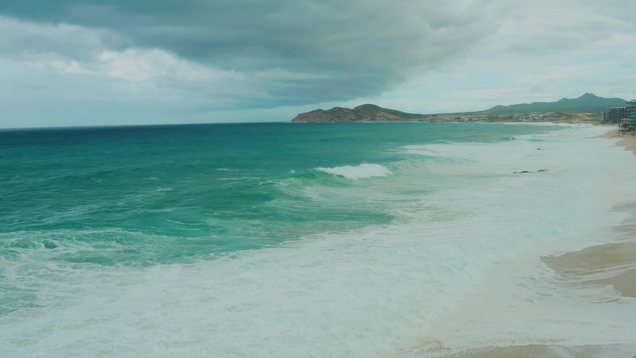 At 0:02, the surf break is more defined in the midground, with white foam curling over turquoise water, pale sand at the right edge, cloudy sky above, and no visible people.