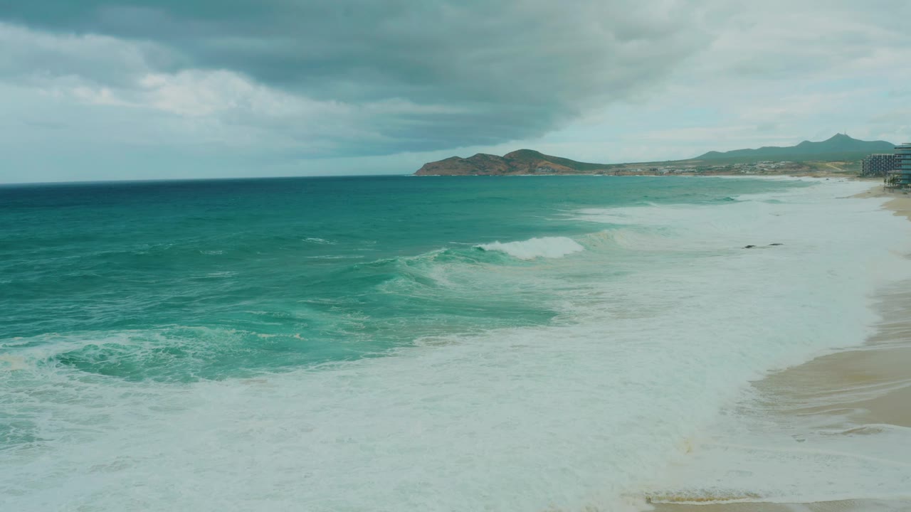 At 0:03, the frame holds a wide aerial view of Dreams Resort Los Cabos surf, with darker turquoise water at left, foam across the beach shallows, distant land, and no visible people.