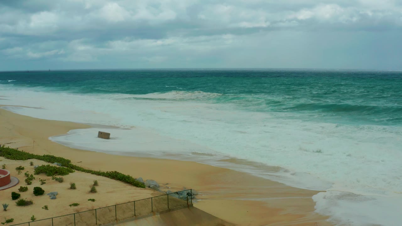 At 0:01, the Dreams Resort Los Cabos shoreline remains framed from above, with white surf spreading over tan sand, low vegetation and fence sections still visible, and no visible people.