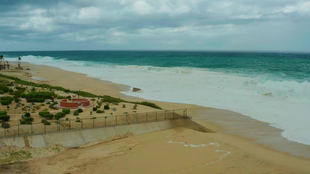 Opening frame at Dreams Resort Los Cabos shows white ocean foam spreading across tan sand beside low greenery, a fence, a red circular beach structure, and no visible people.