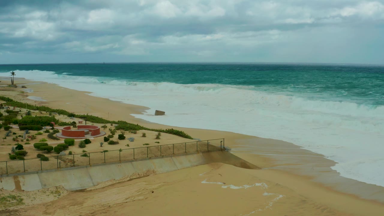 At 0:02, the foam spreads farther along the Dreams Resort Los Cabos beach while the red circular structure and fenced greenery sit at the left edge, with no visible people.